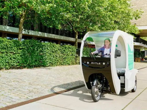 Eine Frau fährt ein Rytle Movr3, ein dreirädriges Elektrofahrzeug, vorbei an einer grünen Hecke und einem Gebäude.