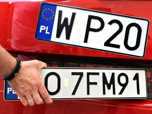 Dłoń z bransoletką montuje nowe, białe tablice rejestracyjne na czerwonym samochodzie. Jedna z nich ma oznaczenie WP20, druga 0-7FM91.