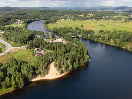 En vacker vy över en svensk landsbygd med en flod som slingrar sig genom gröna fält och skogar. En bro spänner över vattnet, och små röda hus syns bland träden. En idyllisk bild av sommar i Sverige.