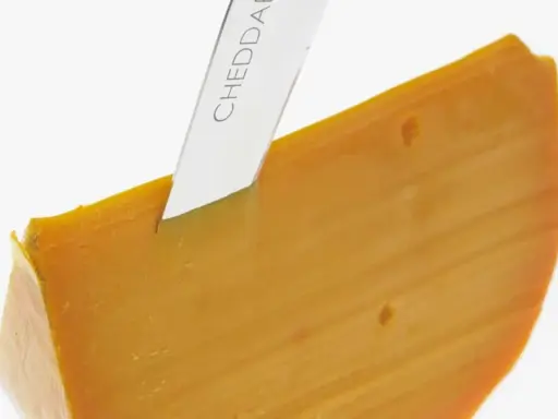 Ein Messer schneidet ein Stück leuchtend orangefarbenen Cheddar-Käse. Was ist Cheddar? Ein köstlicher Hartkäse.
