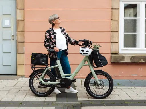 Ältere Dame mit QIO Bike, positive Erfahrungen mit dem E-Bike, das mit einem Helm und einer Tasche beladen ist.