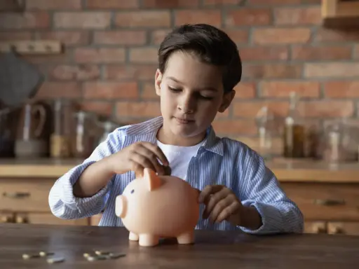 Niño introduce una moneda en una hucha rosa, aprendiendo sobre el ahorro con la cuenta infantil BBVA.