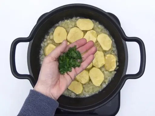Manos añaden perejil picado a patatas en rodajas y cebolla en una sartén, preparando patatas en salsa verde.