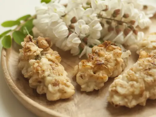 Fiori di acacia fritti, dorati e croccanti, su un piatto di legno, con grappoli di fiori freschi sullo sfondo.