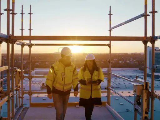 Två byggarbetare i varselkläder inspekterar ett bygge vid solnedgången. De diskuterar säkerhetslösningar, som brandklass EI60.