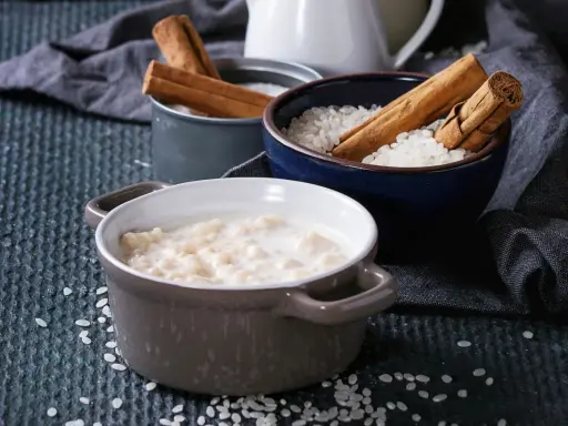 Gotowy do podania ryż na mleku w ceramicznej miseczce, obok miseczki z ryżem i laskami cynamonu.