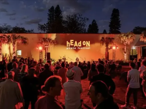Crowd gathered at the hEAd oN photo festival, enjoying a performance under palm trees lit by warm orange lights.