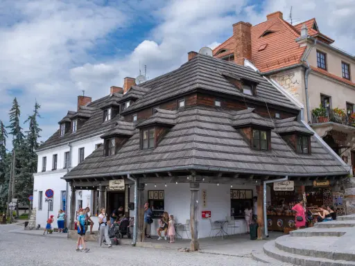 Urokliwy rynek w Kazimierzu Dolnym z charakterystycznymi kamieniczkami i drewnianymi dachami. Ludzie spacerują, kupują lody i pamiątki.