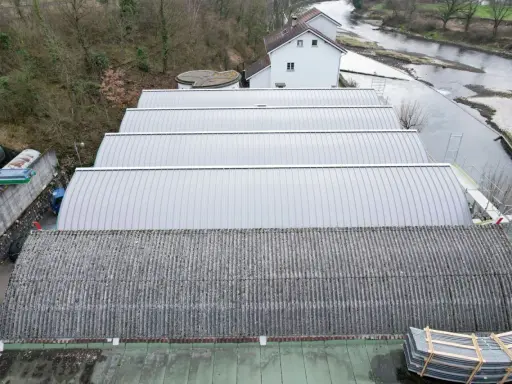 Reihen von Sheddach-Konstruktionen mit Metall- und Wellplatten. Ein Fluss und ein Gebäude sind im Hintergrund zu sehen.