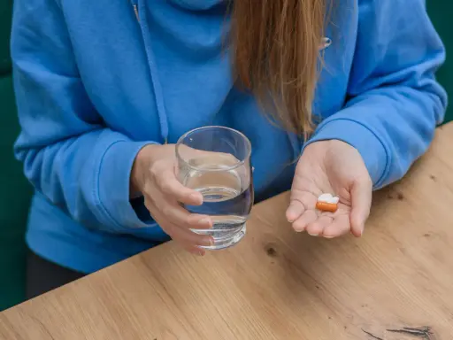Kobieta w niebieskiej bluzie trzyma szklankę wody i kilka tabletek na dłoni.