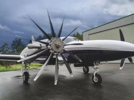 Ein elegantes Flugzeug mit einem beeindruckenden 4-Blatt-Propeller steht auf nassem Asphalt vor einer Halle.