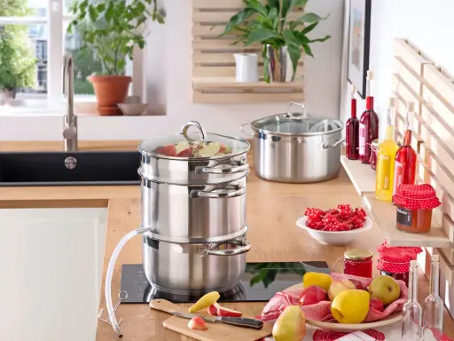 Saft einkochen im Einkochautomat: Obst wird im Dampfgarer zu Saft verarbeitet. Daneben stehen Gläser mit roter Johannisbeermarmelade und Flaschen mit Sirup.