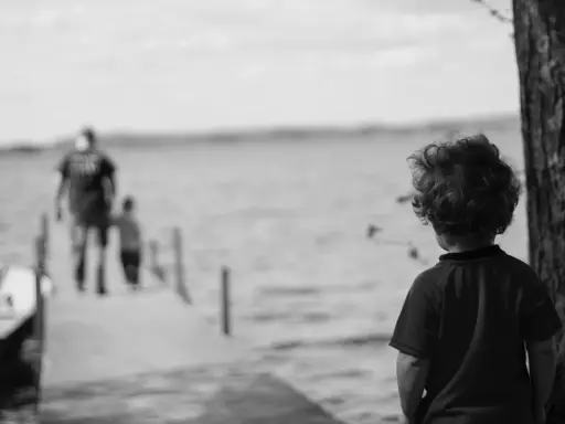 Bambino con capelli ricci guarda verso un molo sfocato dove camminano due figure. Un senso di isolamento, quasi un attaccamento disorganizzato, aleggia nell'aria.