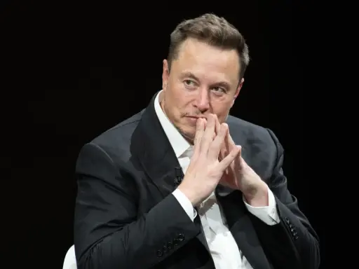 Elon Musk, znany wizjoner i przedsiębiorca, w czarnym garniturze, z dłońmi złożonymi przed twarzą.
