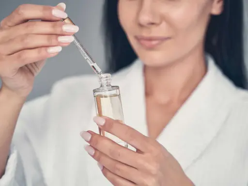 Une femme en peignoir blanc utilise un compte-gouttes pour prélever un sérum, peut-être un mélange de niacinamide et rétinol, pour sa routine de soins.
