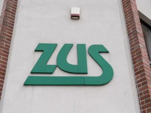 Zielone logo ZUS na białej ścianie budynku z ceglanymi elementami.