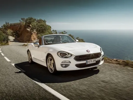 Biały Fiat 124 Spider sunie po krętej drodze nad morzem.