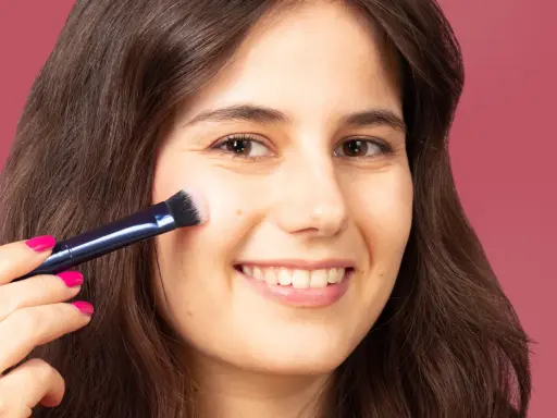 Una mujer sonríe mientras aprende como aplicar colorete con brocha, logrando un rubor natural en sus mejillas.