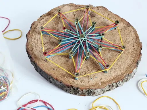 Bunte Gummibänder spannen sich zu einem Stern auf einer Baumscheibe. Ein tolles Bastelprojekt für Kinder, das Kreativität und Feinmotorik fördert.