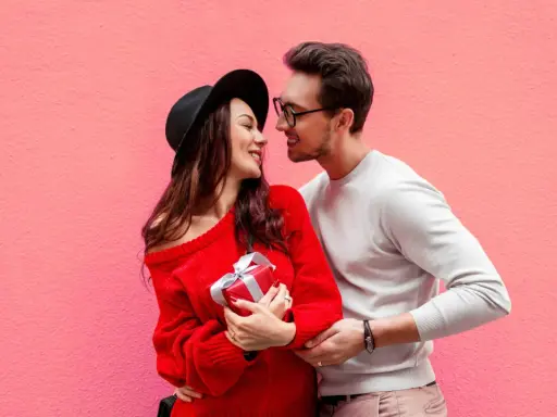 Para zakochanych na tle różowej ściany. Ona trzyma prezent, on obejmuje ją ramieniem. Mówi mu "I love you" po angielsku.