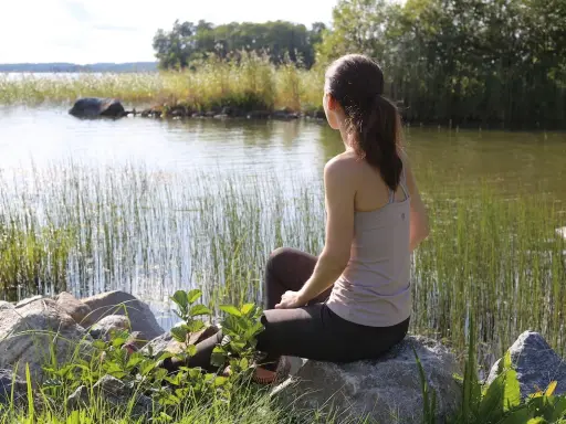 Kvinna sitter vid en sjö och utövar mindfulness svenska. Vassen vajar i vinden, solen glittrar på vattnet.