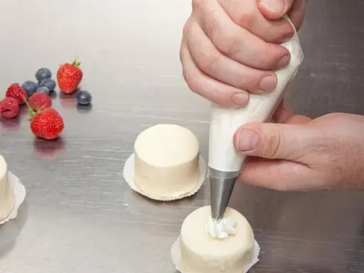 Man usa una manga pastelera para decorar pequeños pasteles. Frutas rojas y arándanos adornan la mesa.