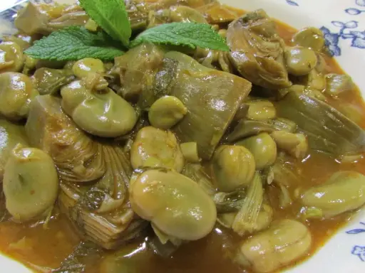 Plato de habas tiernas y trozos de alcachofa en una salsa dorada, decorado con hojas de menta fresca.