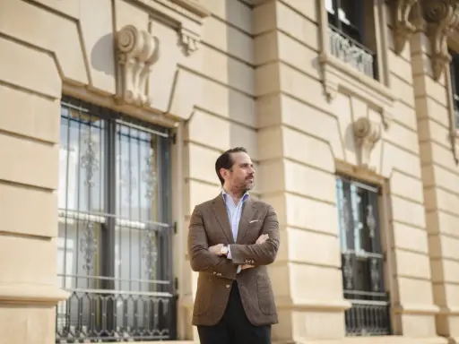 A man in a stylish brown sport coat outfit stands with arms crossed in front of a classic building.