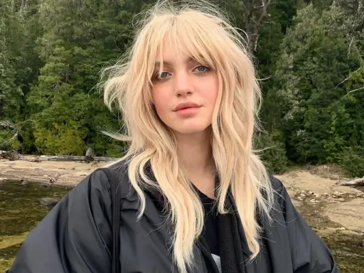 Blonde Frau mit lässigem Wolf Cut stylen, blauen Augen und einem schwarzen Jackett vor einer Waldkulisse.