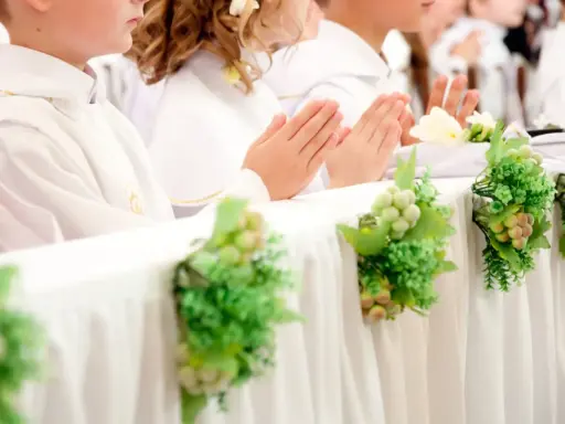 Dzieci w bieli podczas komunii, modlące się ręce. Dekoracje stołu z winogronami i zielenią, symbolizujące koszty komunii w kościele.
