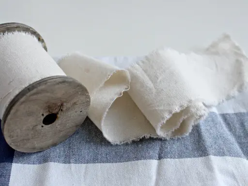 Un vecchio rocchetto di legno con tessuto di cotone pettinato arrotolato. Il tessuto è grezzo e sfrangiato, adagiato su una tovaglia a quadri blu e bianchi.