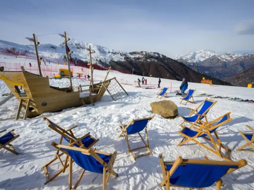 Alpe di Mera: un galeone di legno e sdraio blu attendono i visitatori sulla neve, con montagne innevate sullo sfondo.