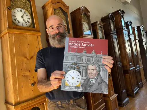 Un homme barbu présente un livre sur Antide Janvier, horloger du roi, devant une rangée de grandes horloges anciennes.