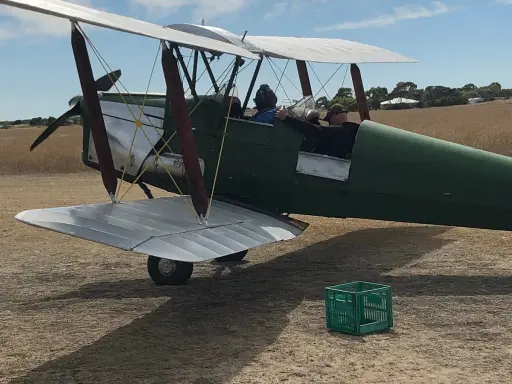 Grüner Doppeldecker, bereit zum Start. Ein Pilot und ein Passagier sitzen im Cockpit. Das Flugzeug erinnert an einen Tiger Moth.