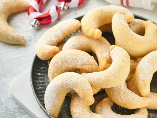Ein Haufen goldbrauner Vanillekipferl, bestäubt mit Puderzucker, liegt auf einem dunklen Teller. Im Hintergrund sind weihnachtliche Zuckerstangen zu sehen.