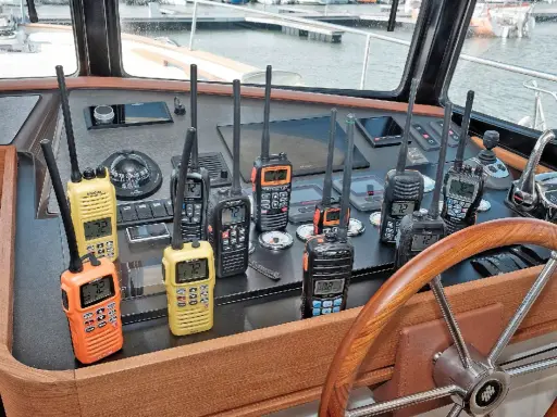 Plusieurs talkies-walkies sur un tableau de bord de bateau. Ils sont de différentes couleurs (orange, jaune, noir) et affichent des canaux. Une discussion sur la différence entre UHF et VHF pourrait être animée avec ces appareils.