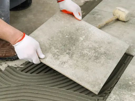 Układanie płytki na kleju w garażu. Ręce w białych rękawiczkach precyzyjnie kładą szarą płytkę na świeżym zaprawie.