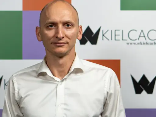 Łysy mężczyzna w białej koszuli na tle kolorowej ściany z logo "wKielcach".