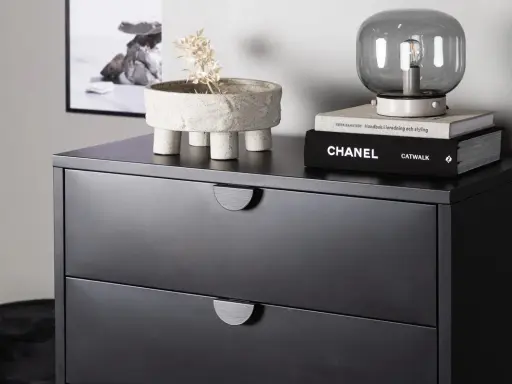 Svart sovrum med en mörk byrå. På byrån står en kruka med torkad växt, en lampa och böcker, bland annat "CHANEL CATWALK".