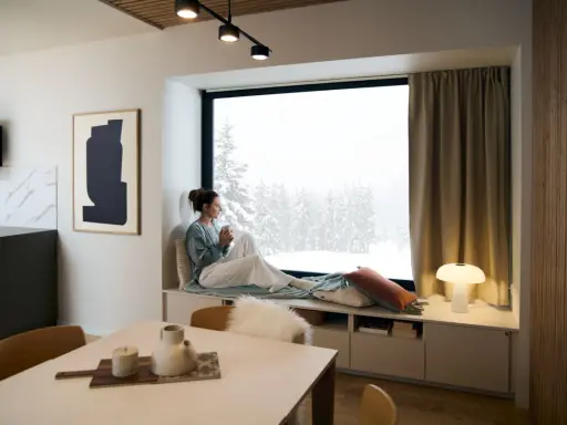 Femme savourant une boisson chaude dans un coin lecture cocooning, regardant la neige tomber.