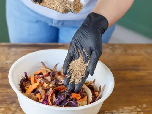 Ajout de bran de riz pour le bokashi maison. Des restes de légumes colorés attendent dans un bol blanc.