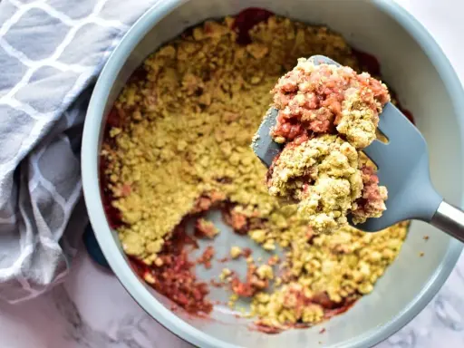 Ein köstlicher Erdbeer Rhabarber Crumble wird mit einem Löffel aus einer Auflaufform gehoben. Daneben liegt ein gemustertes Küchentuch.