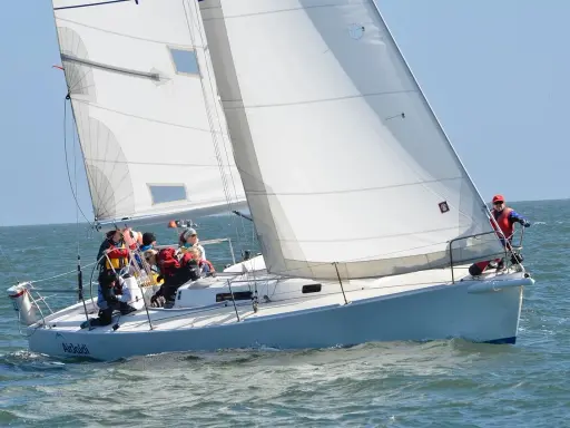 Un voilier blanc navigue sur une mer agitée, avec plusieurs personnes à bord. Le soleil brille, les voiles sont gonflées par le vent.