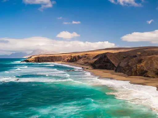 Fuerteventura w lutym: turkusowe fale rozbijają się o piaszczystą plażę, a za nią wznoszą się surowe, brązowe klify.