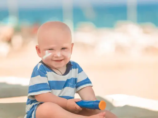 Un bébé souriant avec de la crème solaire sur le visage, tenant un tube de crème solaire. Idéal pour savoir crème solaire bébé à partir de quel âge.