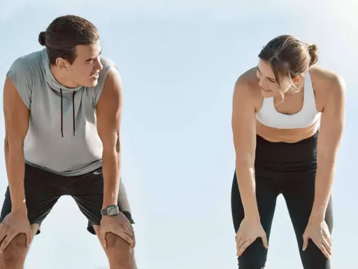 Zwei Läufer erholen sich nach einem anstrengenden 10 km Lauf. Sie schauen sich an, die Hände auf den Knien, erschöpft, aber zufrieden.