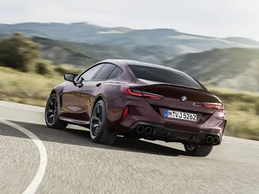 Ein dunkelroter BMW M8 Gran Coupé rast auf einer kurvigen Straße durch eine hügelige Landschaft.
