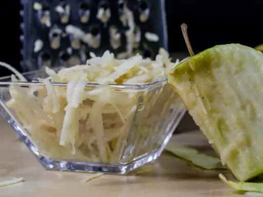 Frisch geriebener Apfel in einer Glasschale, daneben ein Apfelstück und eine Reibe.