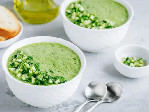 Deux bols de gaspacho concombre frais, garni de dés de concombre et d'herbes. Du pain grillé et de l'huile d'olive à proximité.