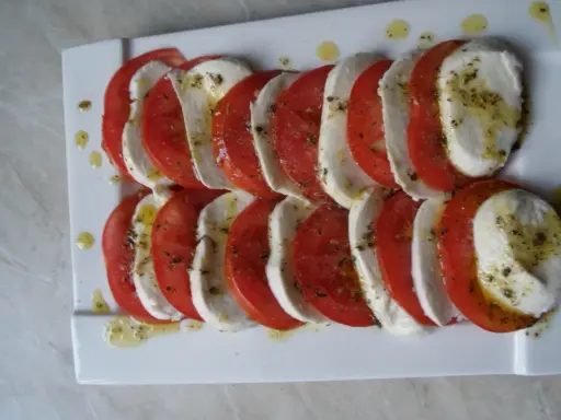 Mozzarella z pomidorami, ułożona naprzemiennie na białym talerzu, skropiona oliwą i ziołami.